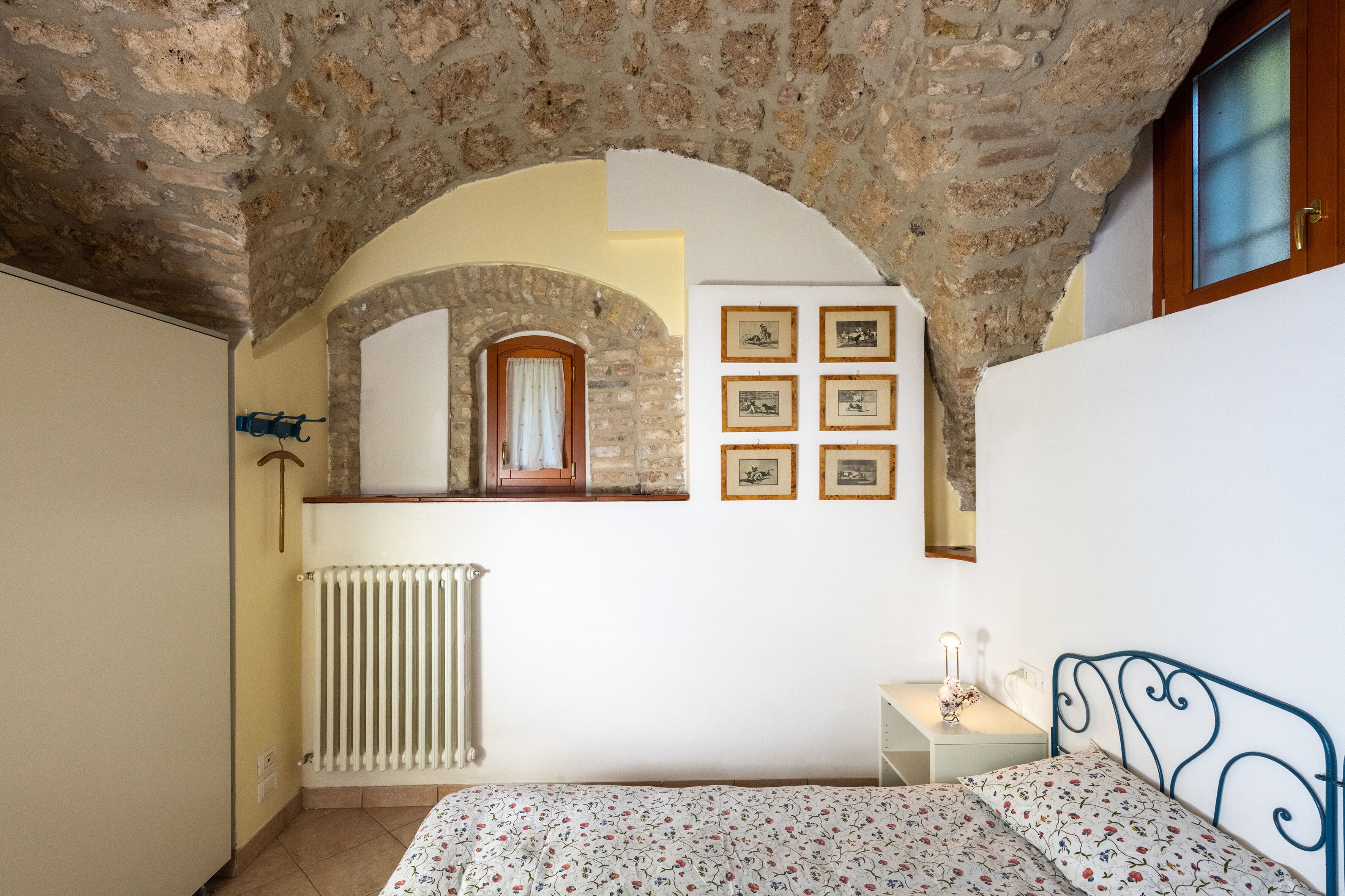 A bedroom with a stone ceiling, a bed with a floral bedspread, a radiator, a wooden door, and framed pictures on the wall.