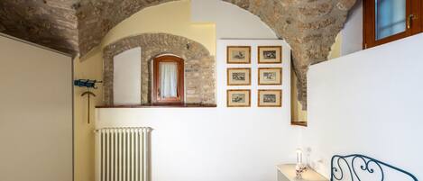 A bedroom with a stone ceiling, a bed with a floral bedspread, a radiator, a wooden door, and framed pictures on the wall.