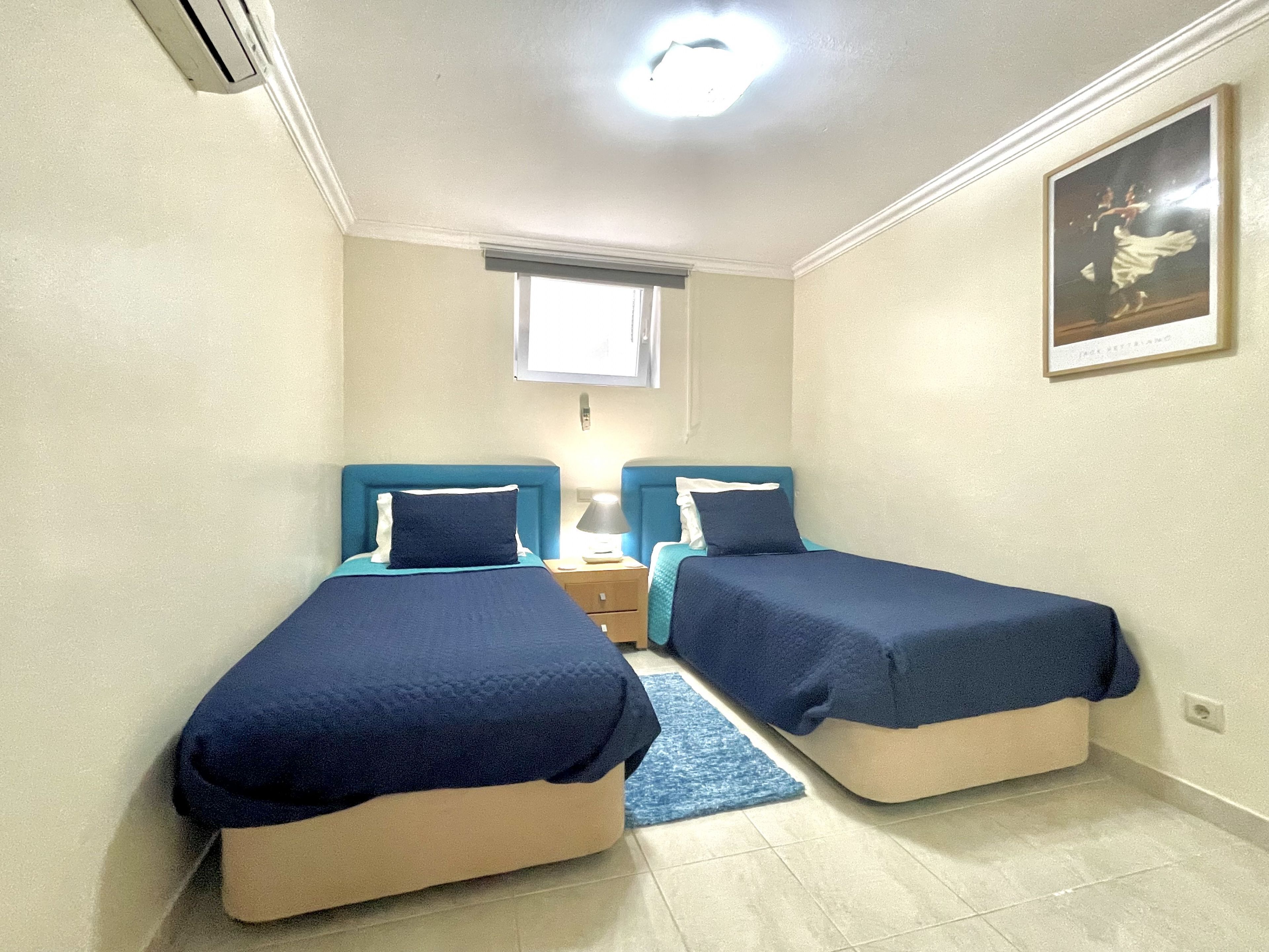 A small, well-lit bedroom with two single beds, a nightstand, a window with blinds, and a framed picture on the wall.