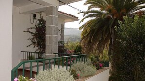 Un balcón con barandilla verde, plantas en macetas y vistas a una palmera y montañas.