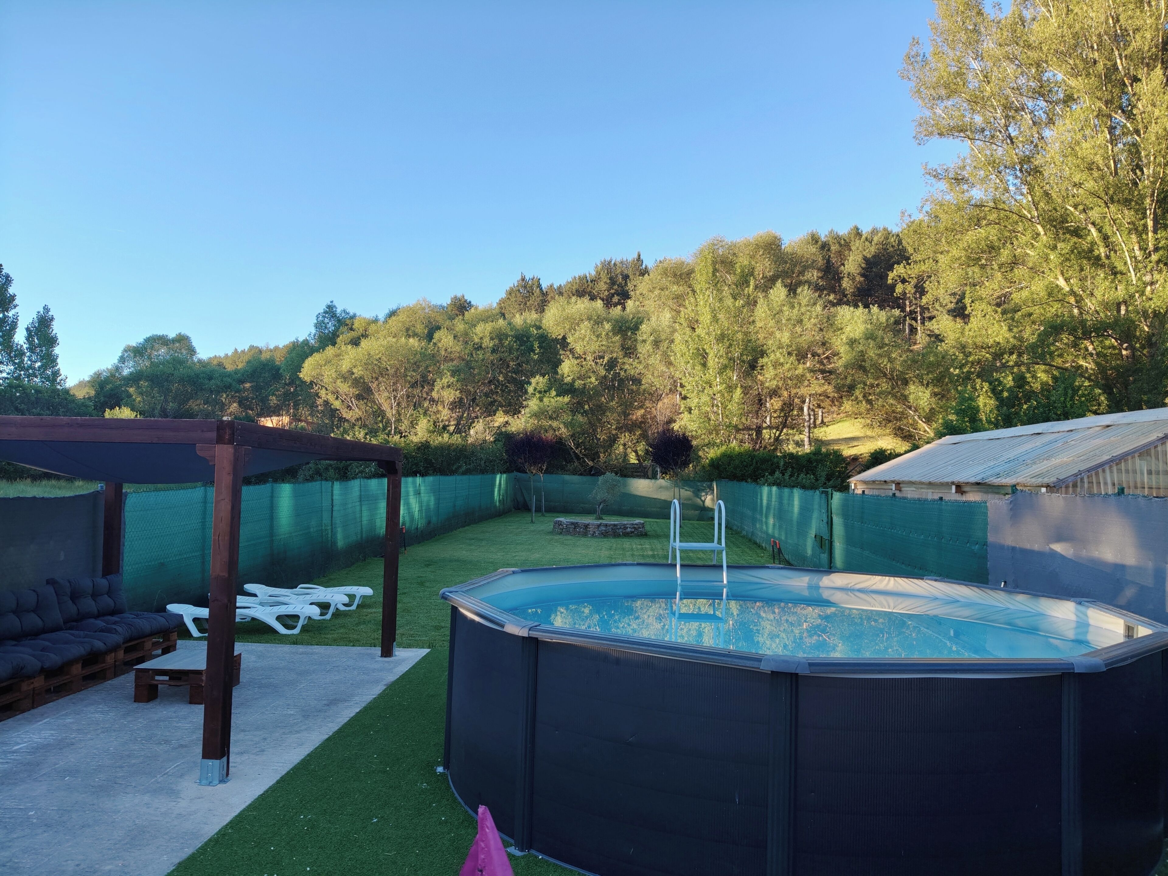 An outdoor swimming pool area with a pergola, lounge chairs, and a fenced garden.