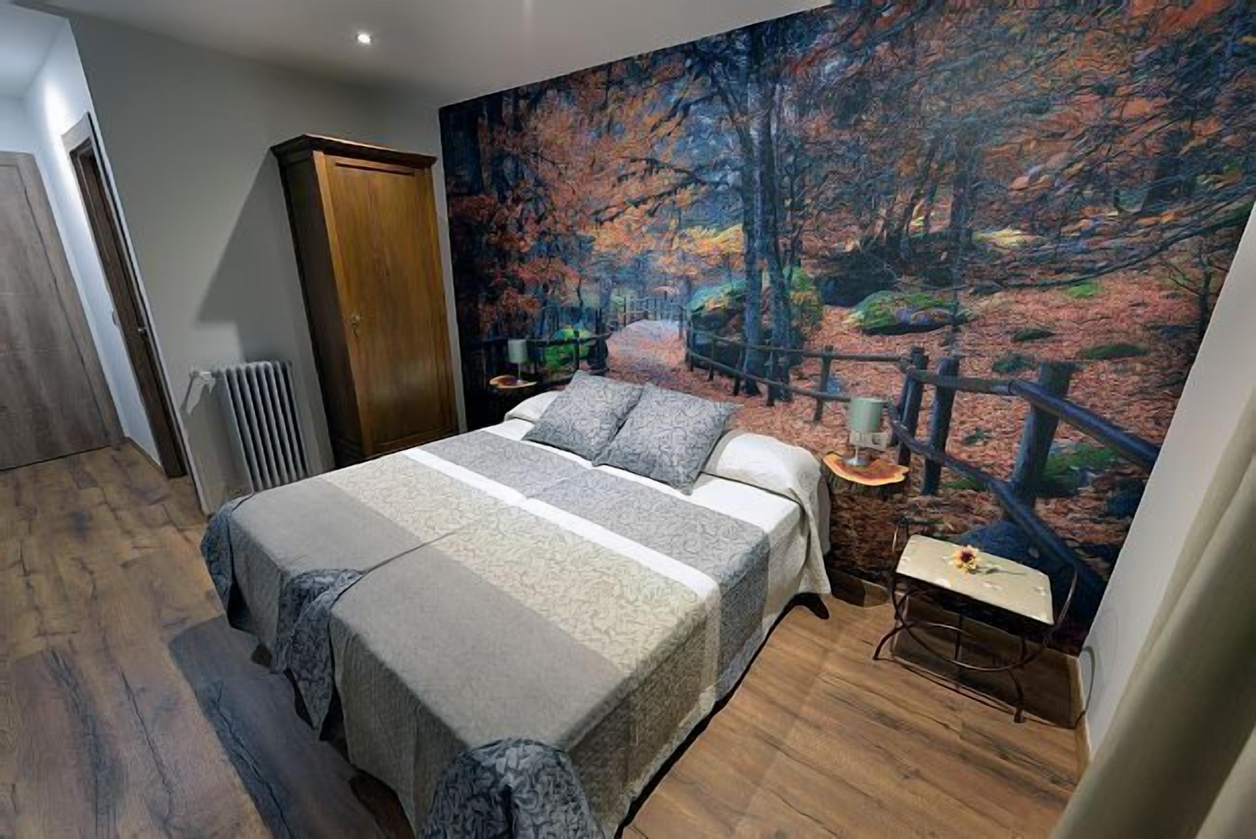 Une chambre avec un grand lit, une armoire en bois, un radiateur, une table ornée d’un vase de fleurs et une fresque murale représentant une scène de forêt.
