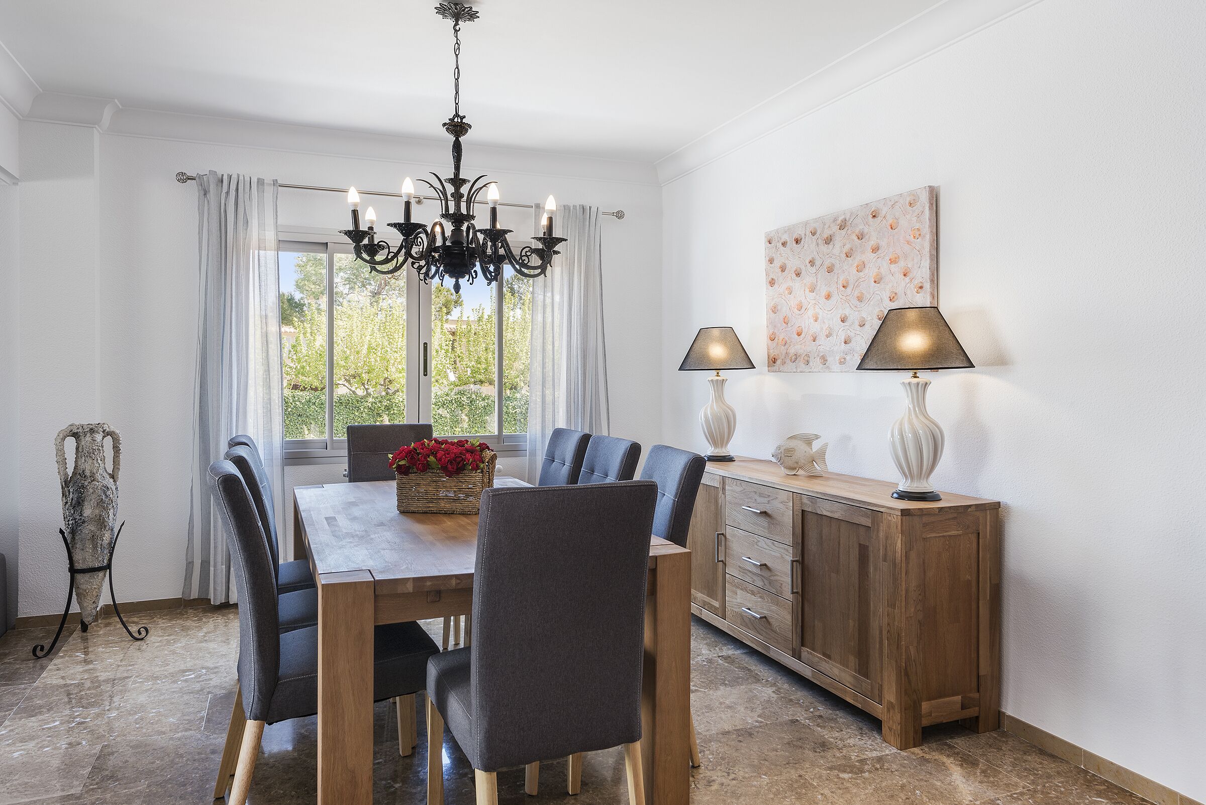 A dining area with a wooden table, grey upholstered chairs, a chandelier, and a cabinet with lamps.
