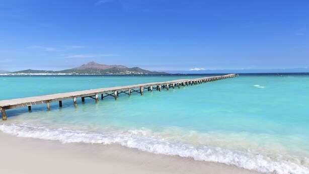 Een houten pier die zich uitstrekt over helder turquoise water, met een zandstrand en een berg op de achtergrond.