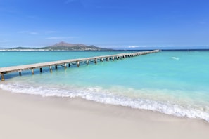 Une jetée en bois s’étendant dans des eaux turquoise et limpides, avec une plage de sable et une montagne en arrière-plan.