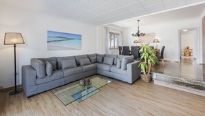 Een moderne woonkamer met een grijze hoekbank, een glazen salontafel, een potplant en een schilderij van een strandgezicht.