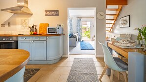 A modern kitchen with a built-in oven, microwave, and a stainless steel range hood. There is a dining area with a wooden table and a chair, and a view of a garden through an open door.