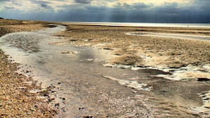 A beach with a curved water channel and a distant view of the sea under a cloudy sky.