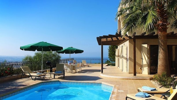 Un espace piscine avec des chaises longues et des parasols, une vue sur la mer et un bâtiment agrémenté d’une pergola.
