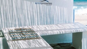 Un espace extérieur aménagé près de la plage, avec une table et un banc, un luminaire suspendu et le logo de « Lemosproperties, agence immobilière ».