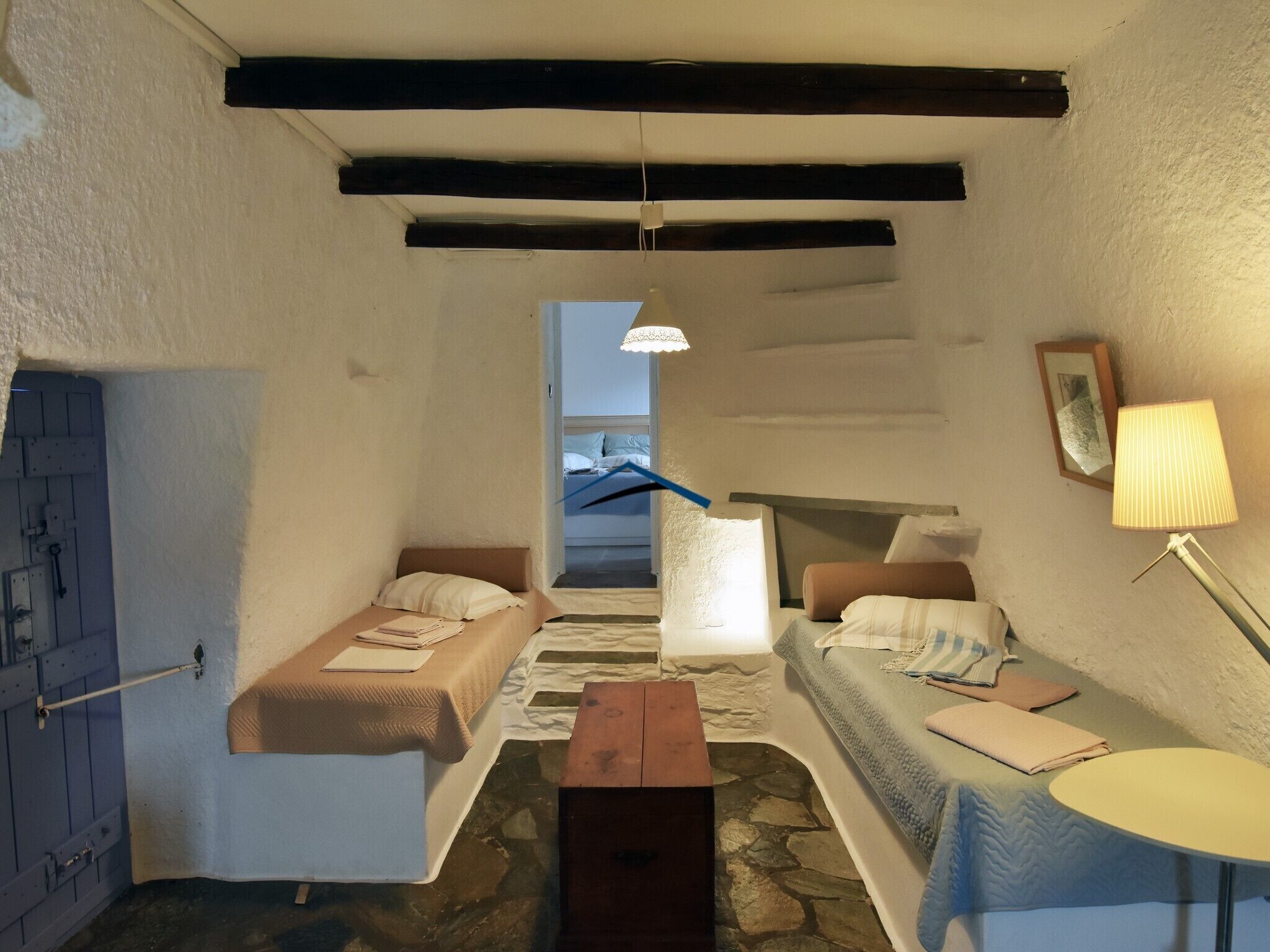 A cozy room with a stone floor, a wooden bench, two beds with pillows, a small table, and a lamp.