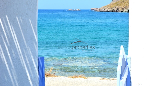 Un arc de pierre mène à une plage bordée d’une eau d’un bleu limpide, et on aperçoit au loin un promontoire rocheux.