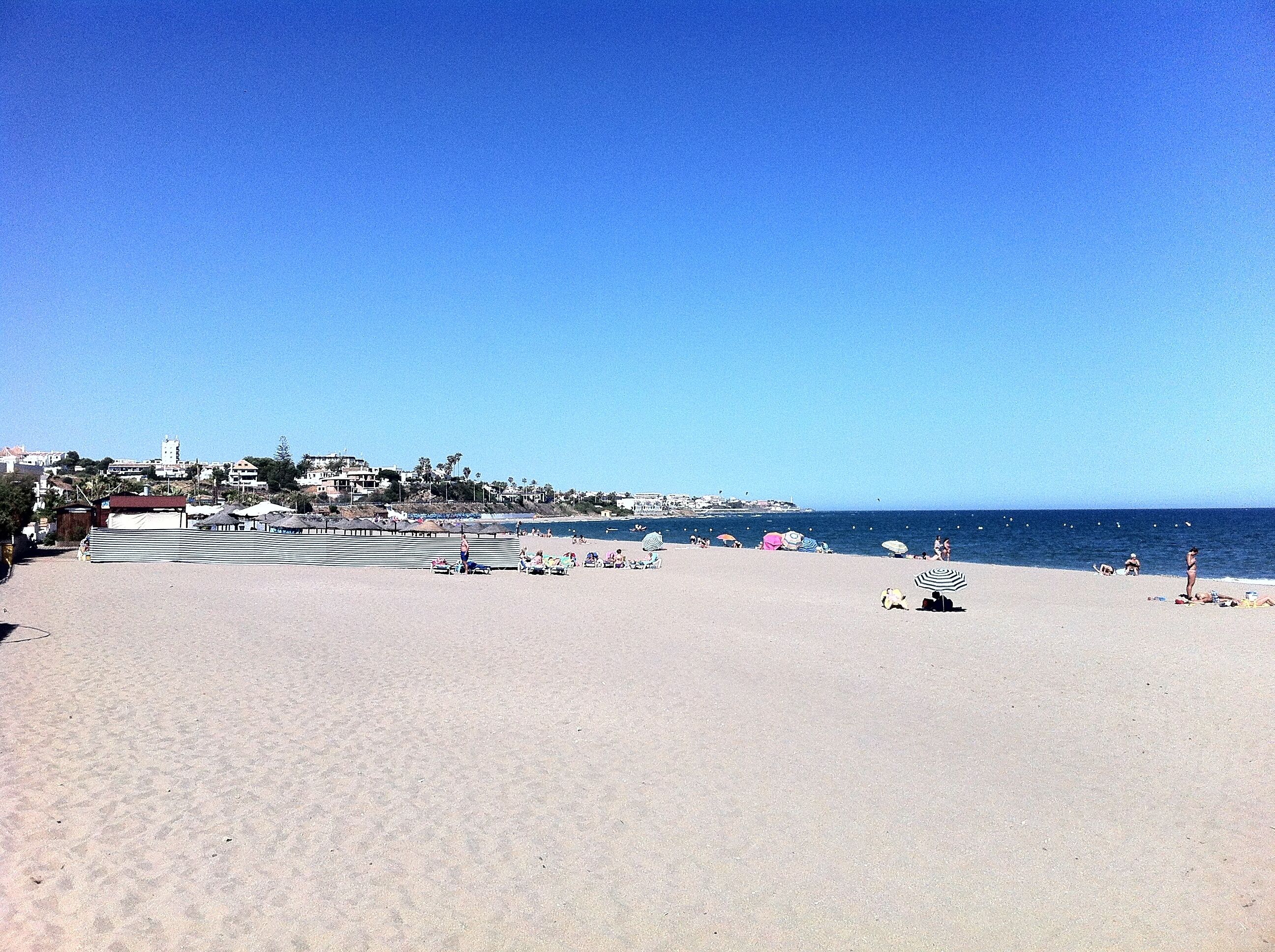Ein Strand mit blauem Himmel, einigen Sonnenschirmen und einer Stadt in der Ferne.