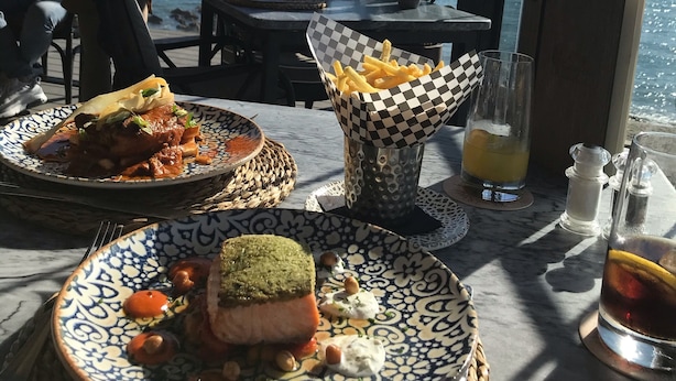 A dining table with plates of food, a glass of drink, and a view of the sea. - EASY WALK TO MAX BEACH LOVELY APT  WI-FI  STUNNING SEA/POOL VIEWS +ELEVATOR/LIFT (Riviera del so/l La cala)