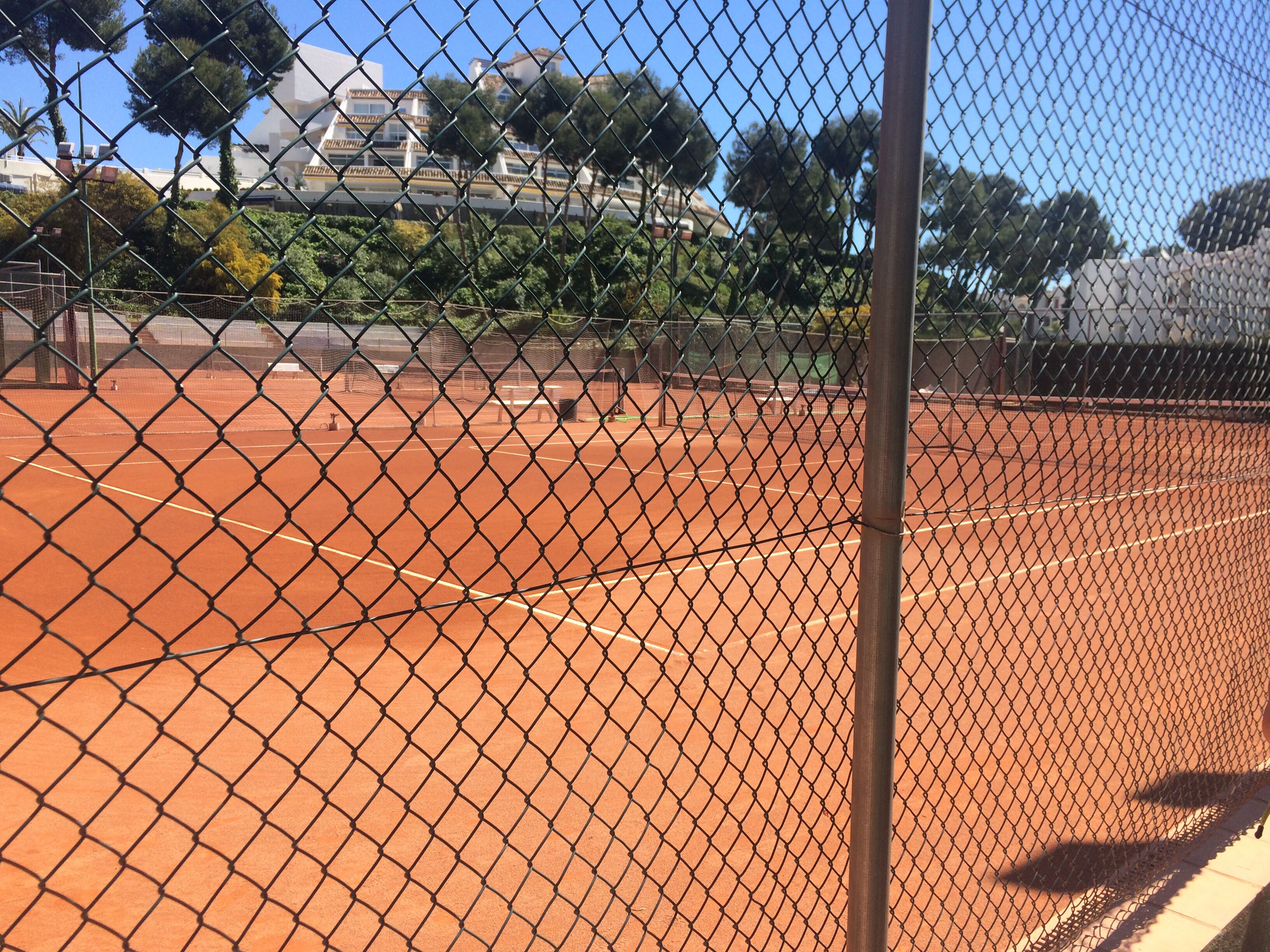 Tenniskenttä, jonka ympärillä on metallinen verkkoaita, ja taustalla näkyy kirkas taivas.