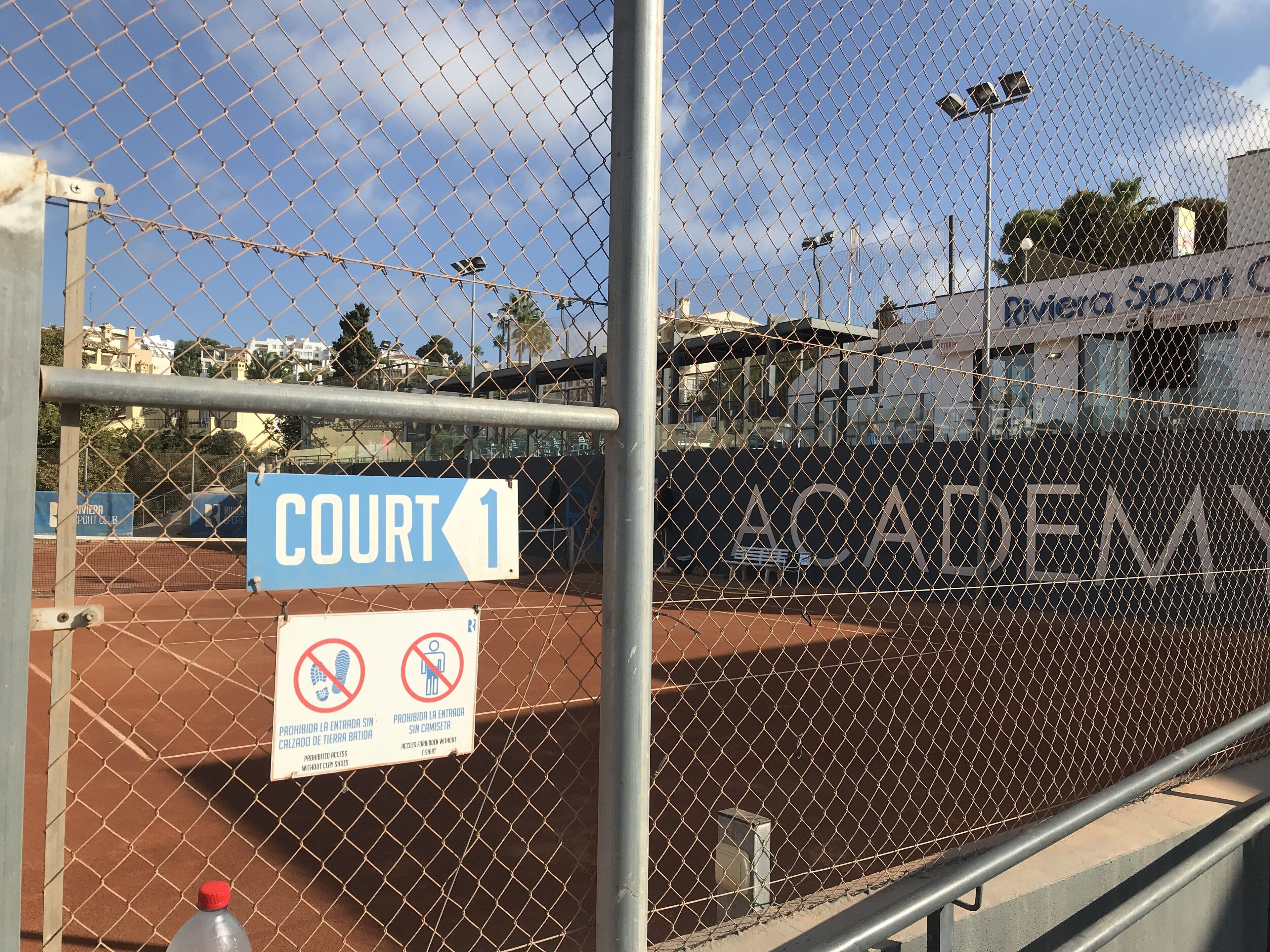 A tennis court with a chain-link fence, a sign indicating 'COURT 1', and a no-entry sign for unauthorized access.