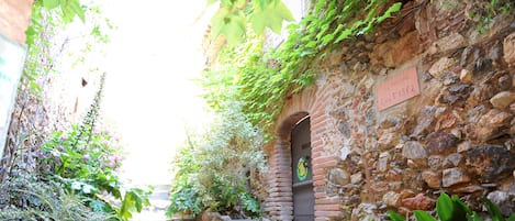 A narrow stone pathway flanked by lush greenery and a stone wall with a door and a sign.
