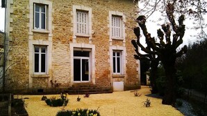Ein Gebäude aus Stein mit einem zentralen Eingang, zahlreichen Fenstern und einem Garten mit gelben Blumen und einem Baum.