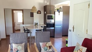 A spacious living area with a wooden ceiling, grey armchairs with decorative pillows, a red sofa, and a wooden coffee table. A ceiling fan is visible, and the room leads to a kitchen with modern appliances.