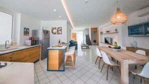 Une cuisine moderne dotée d’un îlot central, d’un coin repas avec une table en bois et des chaises blanches, et d’un escalier menant à un niveau supérieur.