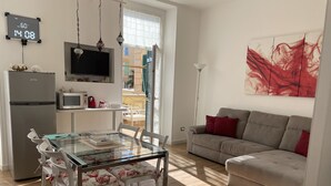 Un salon moderne avec une table à manger en verre, des chaises blanches, un canapé beige agrémenté de coussins rouges, un téléviseur à écran plat, un réfrigérateur et un coin cuisine équipé d’un four à micro-ondes.