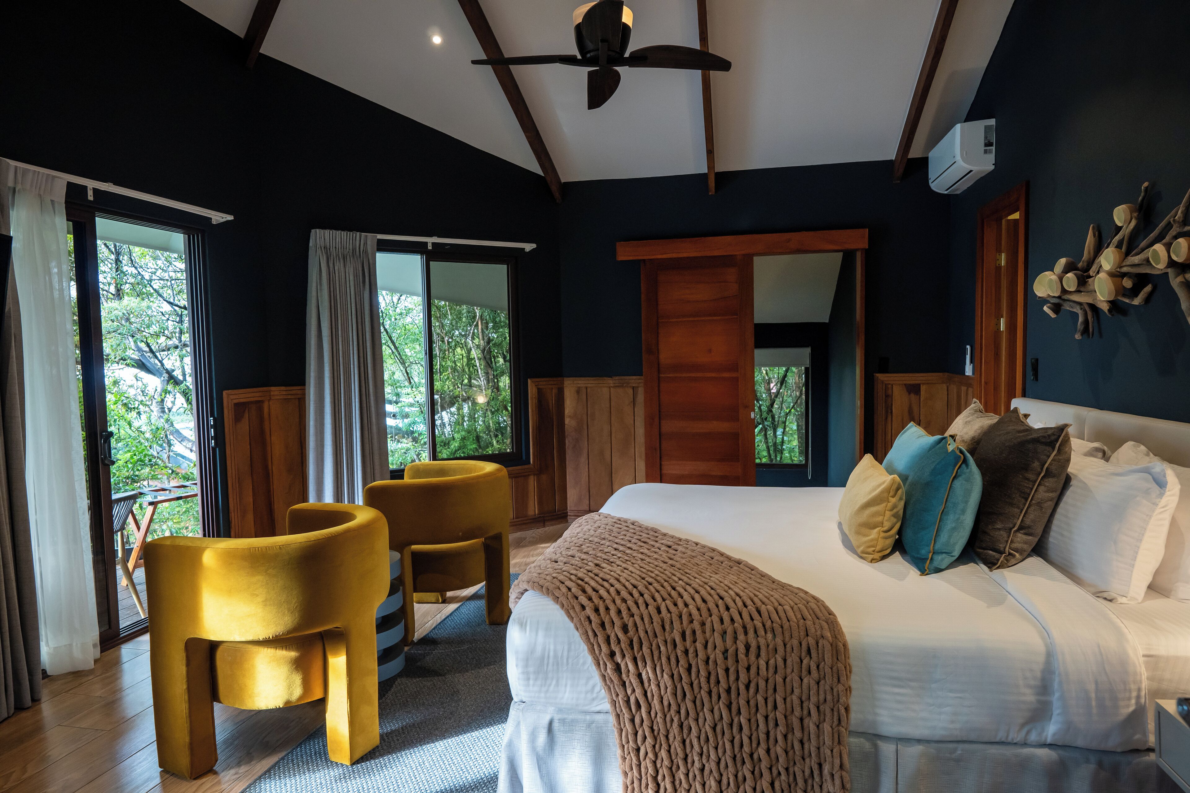 A bedroom with a large bed, a ceiling fan, and a view of trees through the windows.