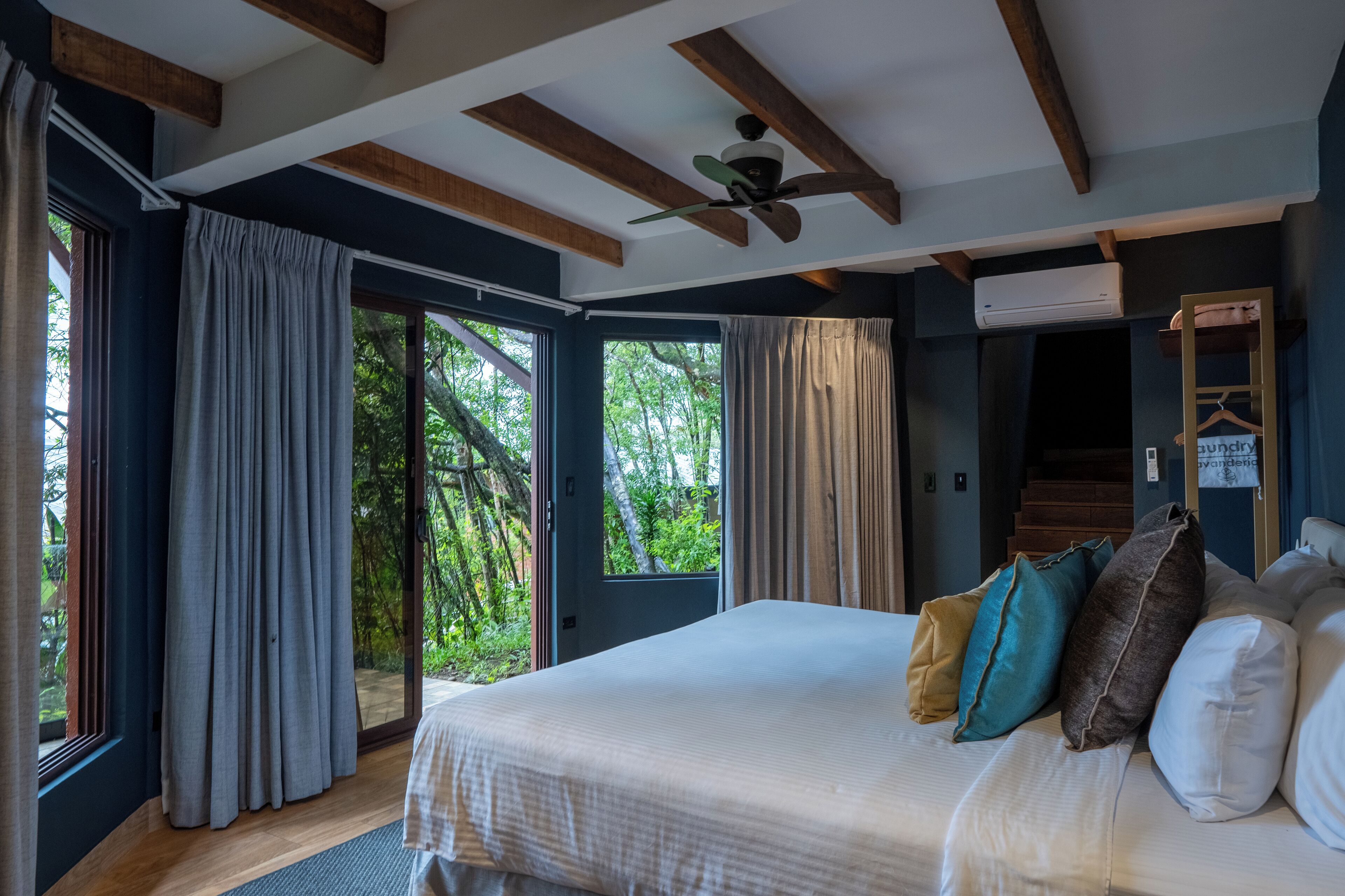 Camera da letto con un letto grande, numerosi cuscini, una ventola a soffitto e vista sul verde dalle finestre.