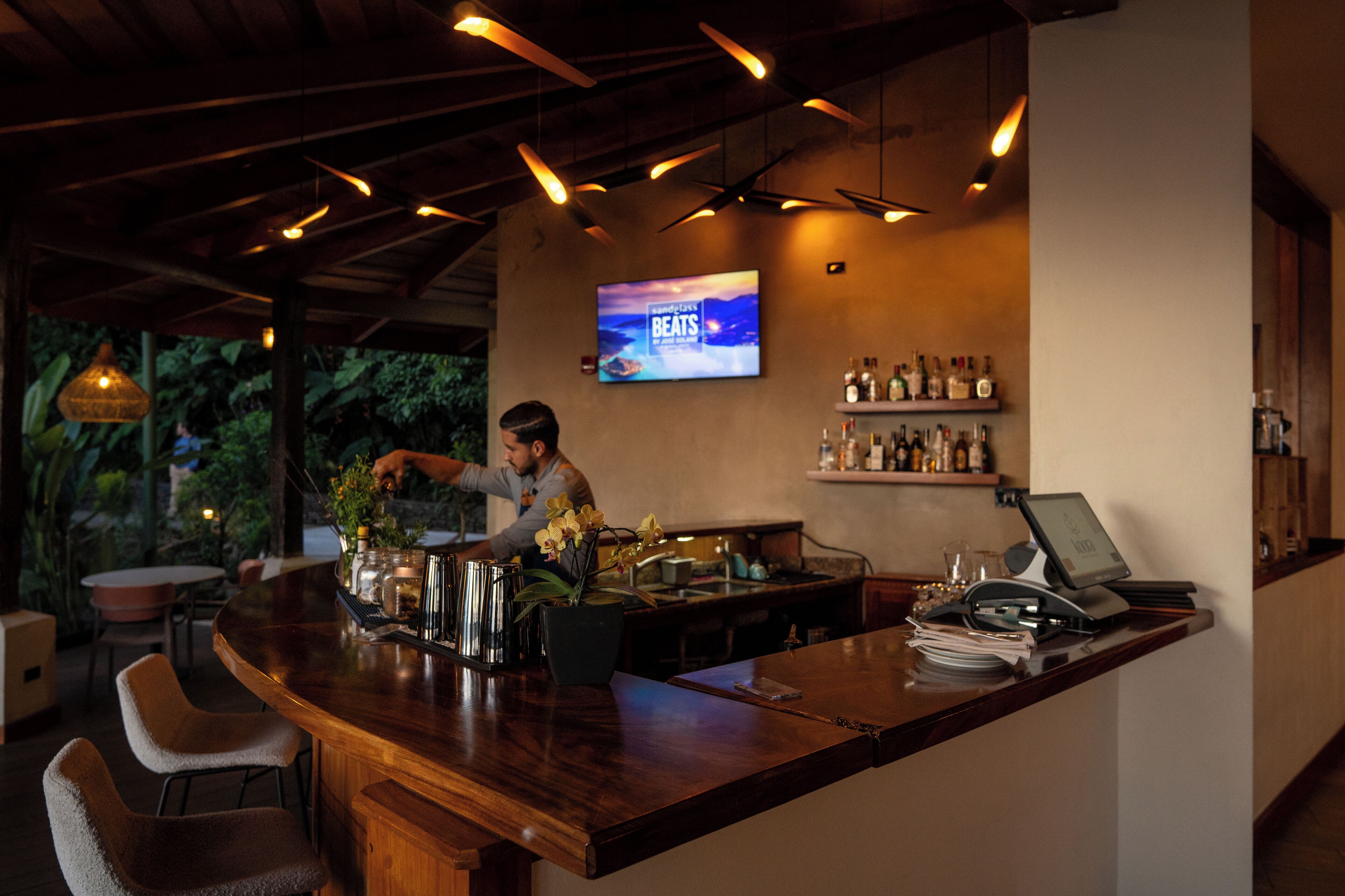 A bar area with a wooden counter, bar stools, a television screen, and shelves with bottles.