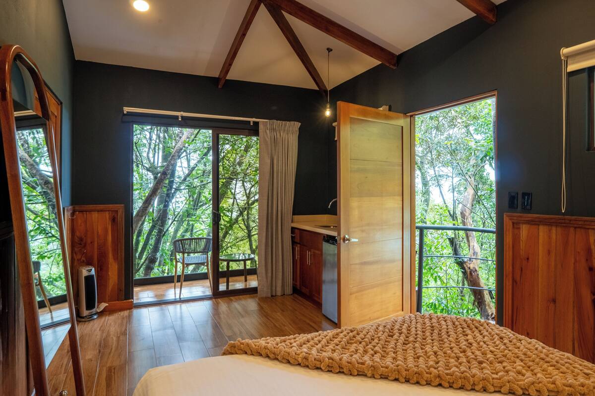 A bedroom with a large bed, a wooden desk, and a balcony with a view of trees.