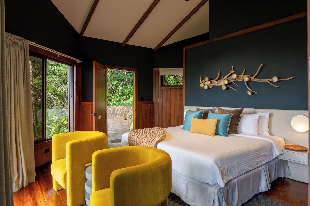 A bedroom with a large bed, yellow chairs, a wooden ceiling, and a view of greenery outside.
