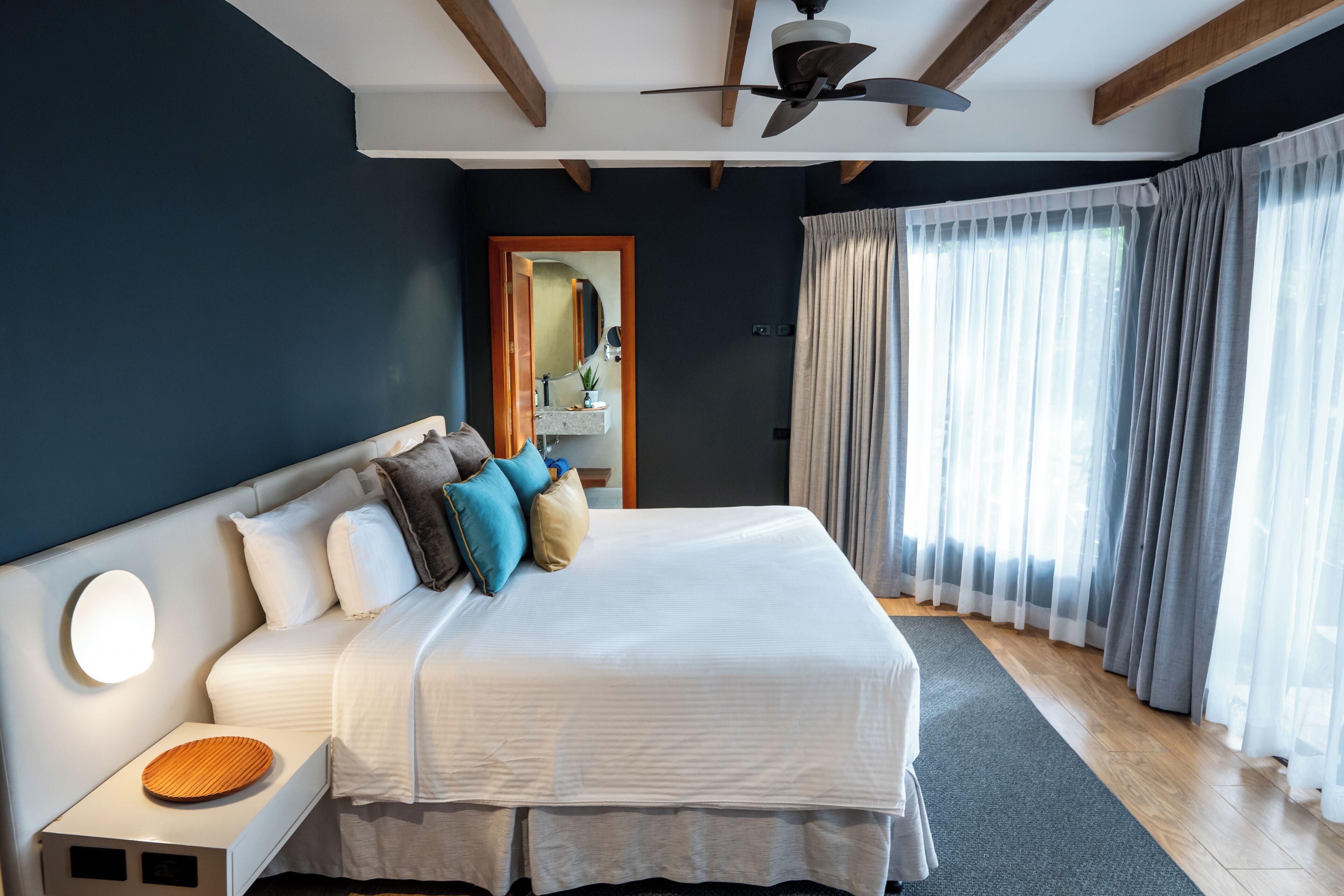 A modern bedroom with a large bed, a nightstand, a ceiling fan, and a view of the outdoors through sheer curtains.