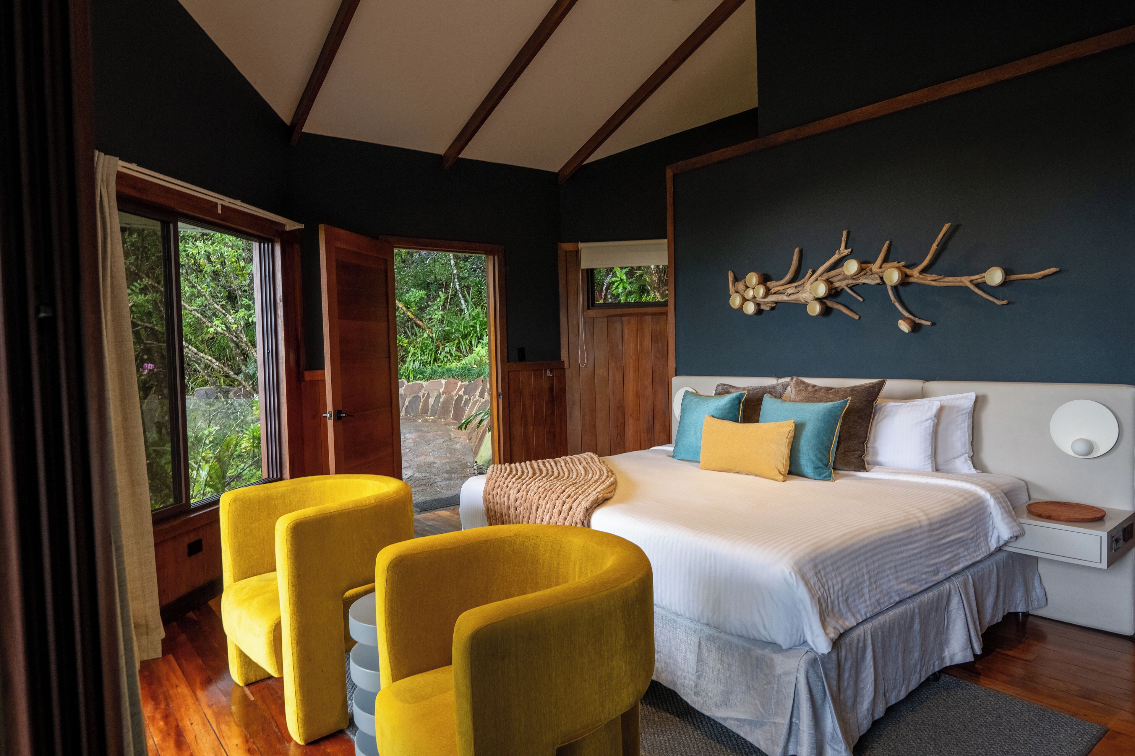 A bedroom with a large bed, two yellow chairs, a wooden headboard, and a wall-mounted light fixture.