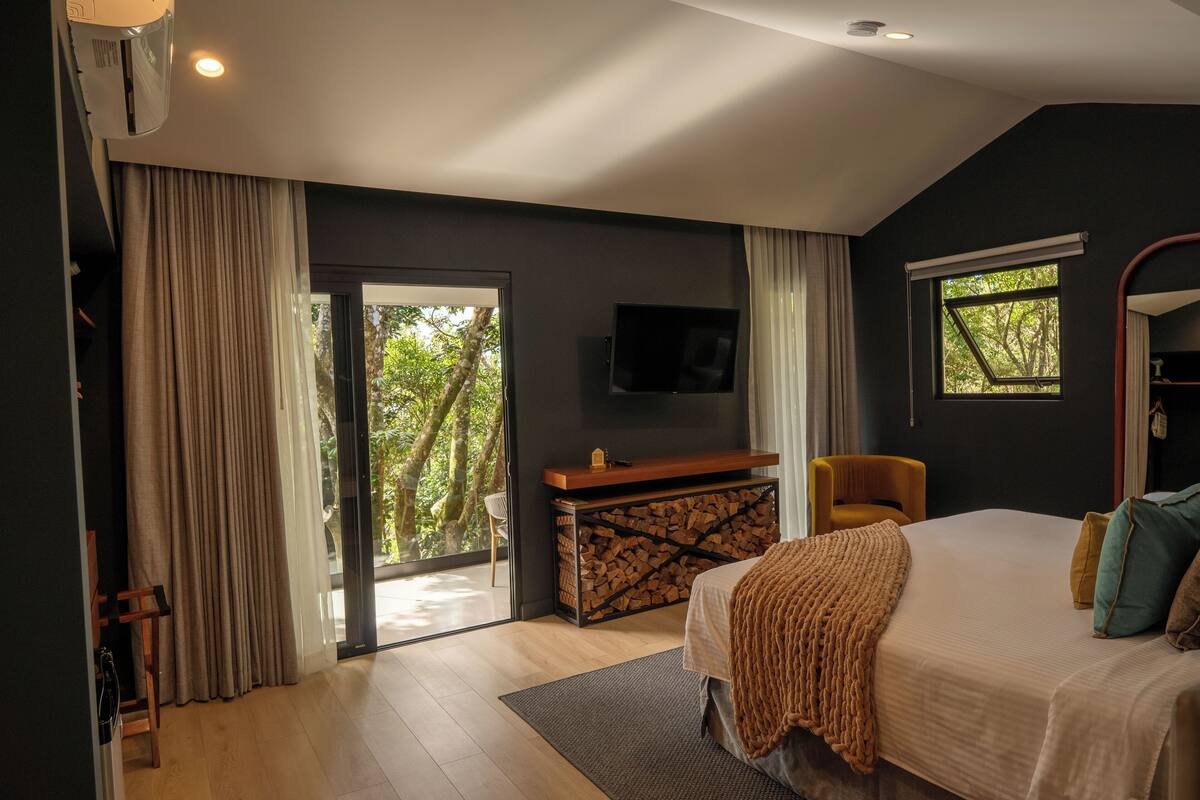 A bedroom with a bed, a TV, a desk with a chair, and a window with a view of trees.