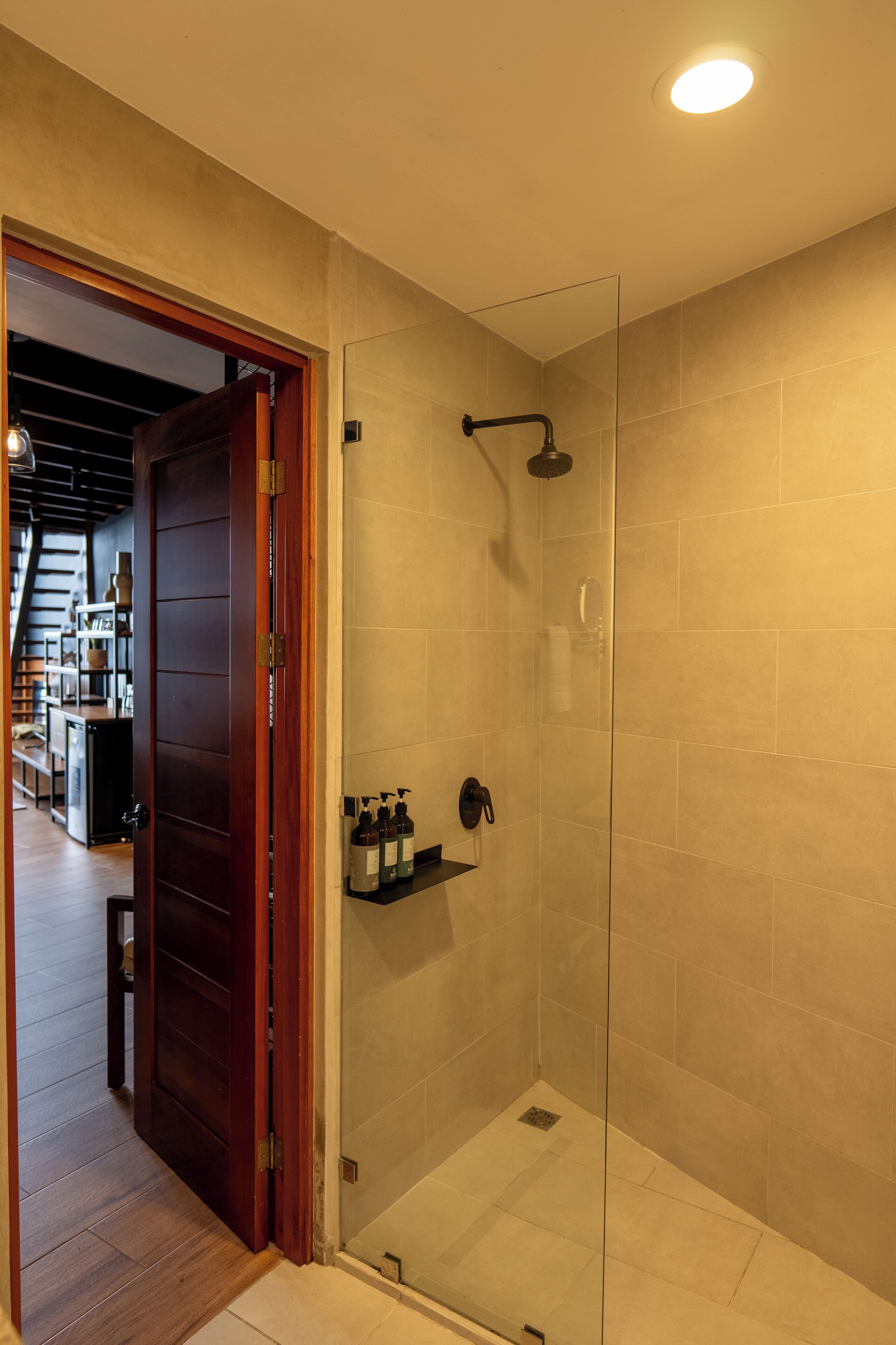 Une douche fermée par des parois de verre, avec des carreaux de couleur beige, une porte en bois et une étagère contenant des articles de toilette.
