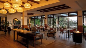 A spacious indoor area with multiple tables and chairs, large windows offering a view of greenery outside, and hanging wicker lamps providing warm lighting. - Koora Hotel by Sandglass (Monteverde)