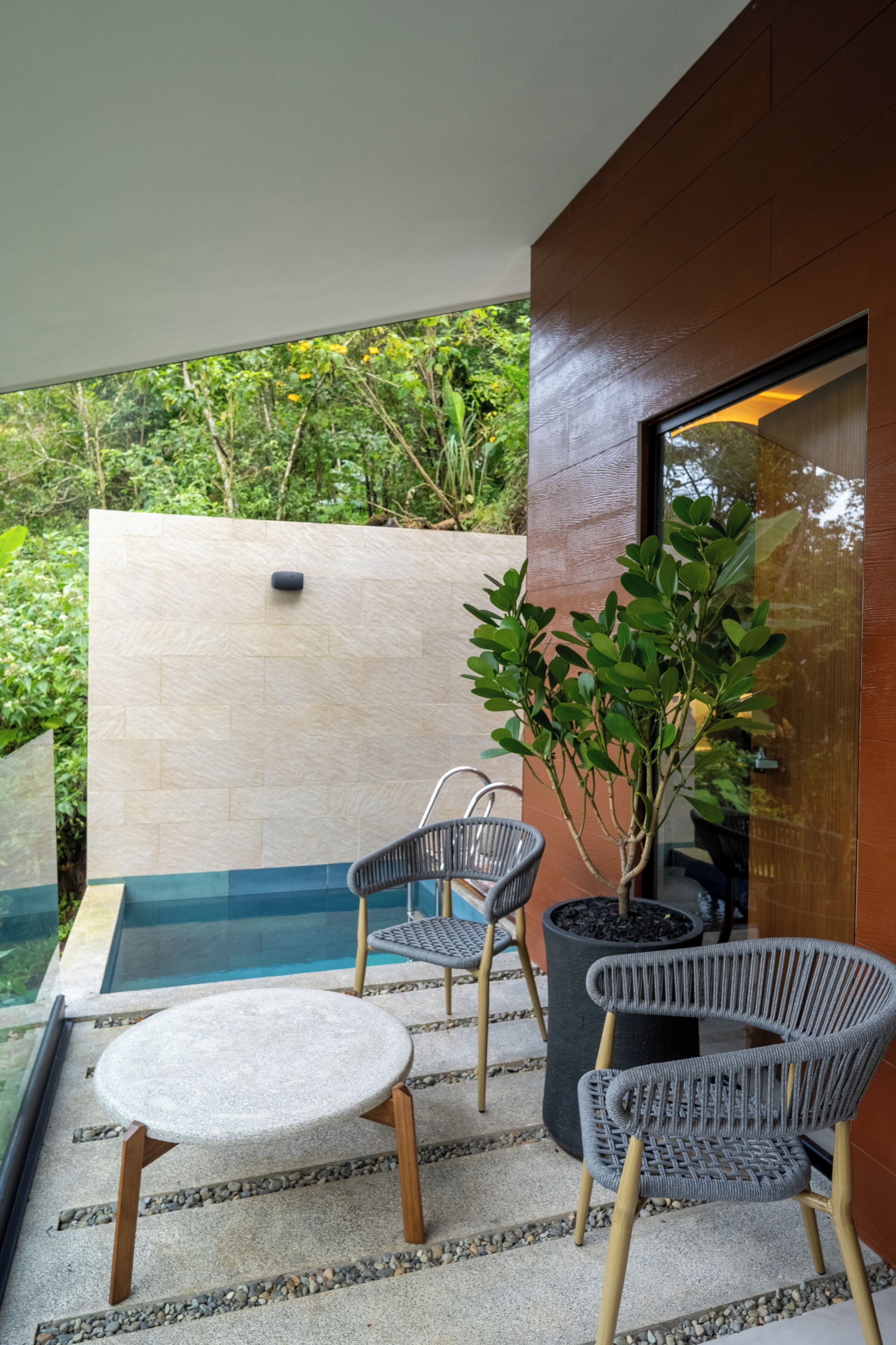 A modern outdoor seating area with a round table and two wicker chairs, a potted plant, and a view of a swimming pool and lush greenery.