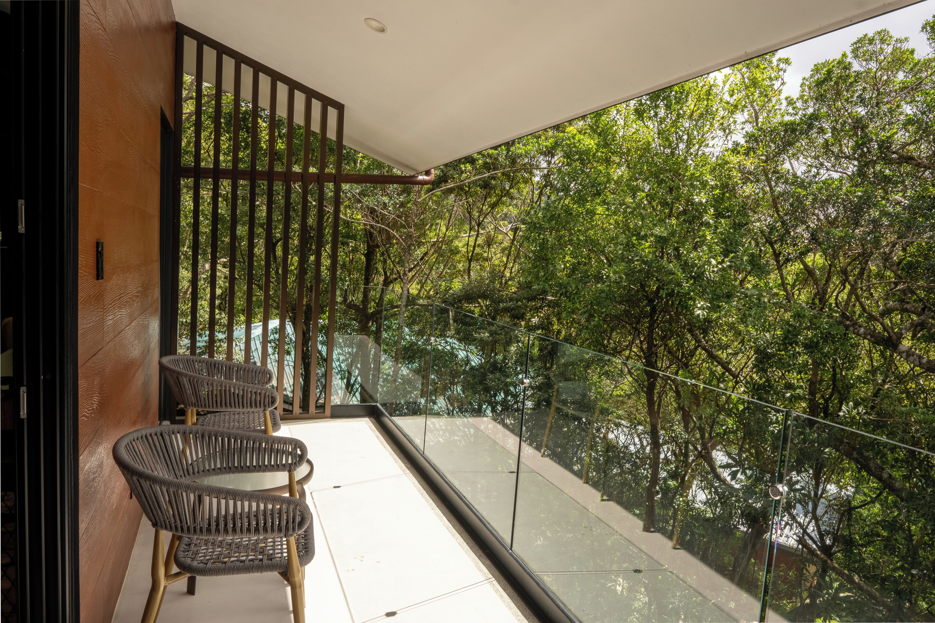 Een balkon met rieten stoelen, een glazen balustrade en uitzicht op dichte bomen.