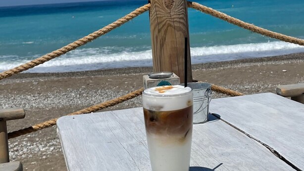 Un vaso de café helado sobre una mesa de madera junto a la playa, con una barrera de cuerda y un poste de madera al fondo.