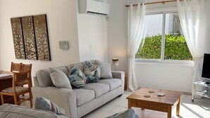 Una sala de estar con un sofá gris, una mesa de centro de madera, un mueble para televisión, un ventilador de techo y una ventana con cortinas.