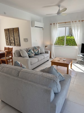 Ein Wohnzimmer mit grauem Sofa, einem Holztisch, einer TV-Konsole, einem Deckenventilator und einem Fenster mit Vorhängen.
