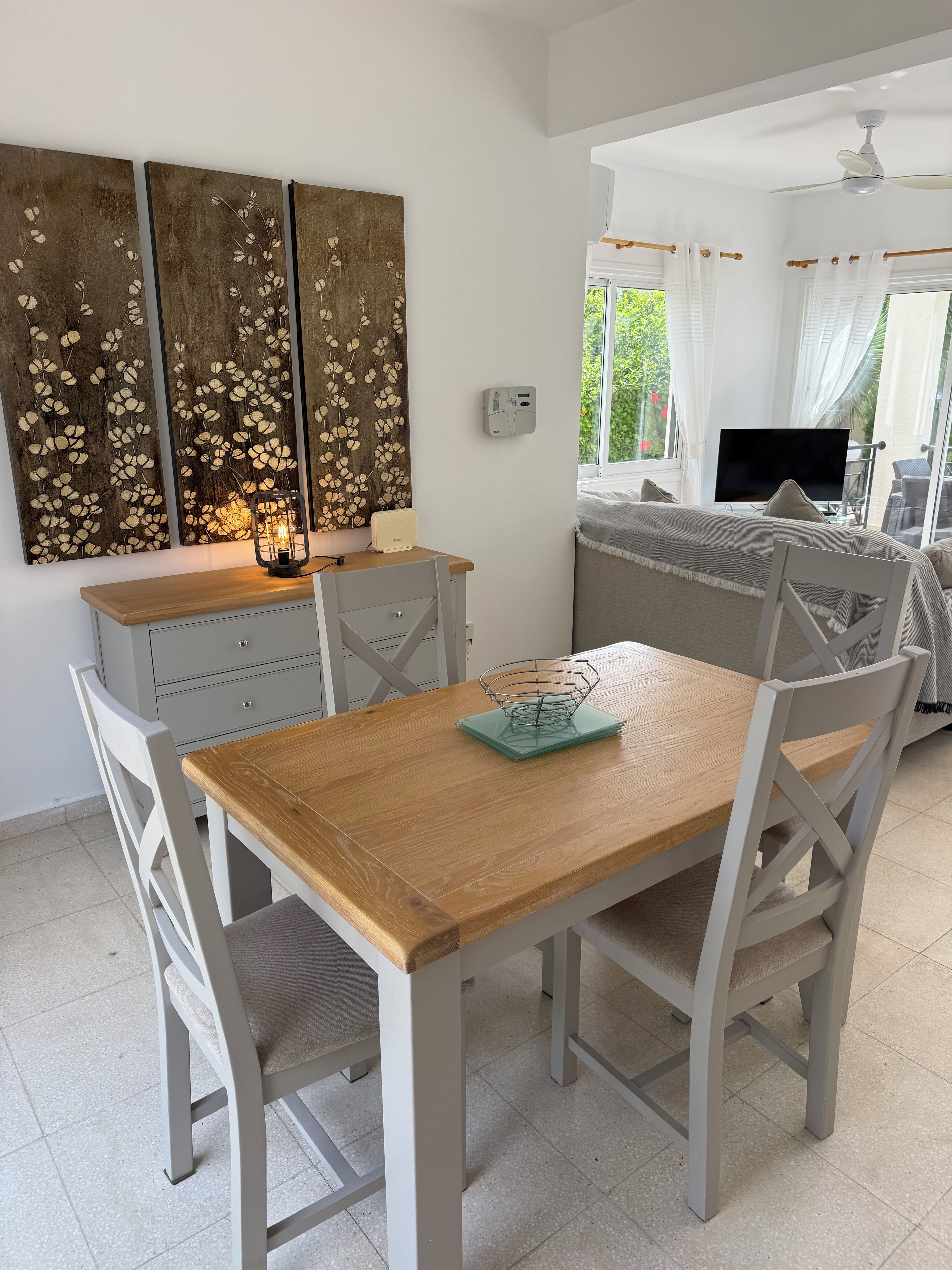 A dining area with a wooden table and white chairs, a TV, and a view of greenery outside.