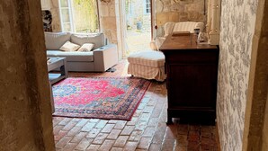 Una stanza con pavimento in mattoni, un tappeto decorato, una poltrona, un mobile in legno e una finestra vista verde.