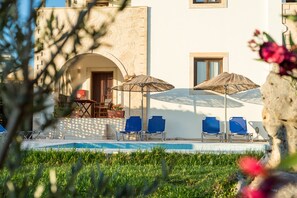 A poolside area with blue lounge chairs and two umbrellas, a stone building with an arched entrance, and a potted plant.