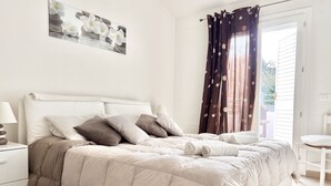 A neatly made bed with a beige duvet, a bedside table with a lamp, and a window with curtains. - Villa Etienne - Key to Villas (Valledoria)
