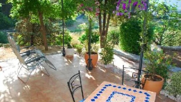 An outdoor patio with a tiled table and chairs, surrounded by potted plants and trees.