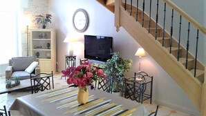 Una sala de estar con una escalera, un televisor, una mesa de comedor con flores y una estantería.