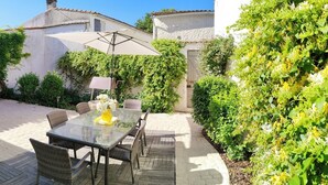 Un patio al aire libre con una mesa de vidrio y sillas de mimbre, rodeado de abundante follaje y un edificio blanco.