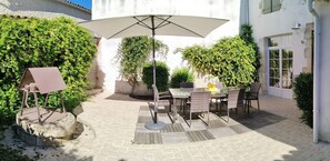 Een terras buiten met een tafel en stoelen, omringd door groen en een wit gebouw.