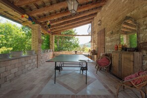 Une terrasse couverte avec une table de ping-pong, des fauteuils en osier, un meuble en bois et un mur en briques.