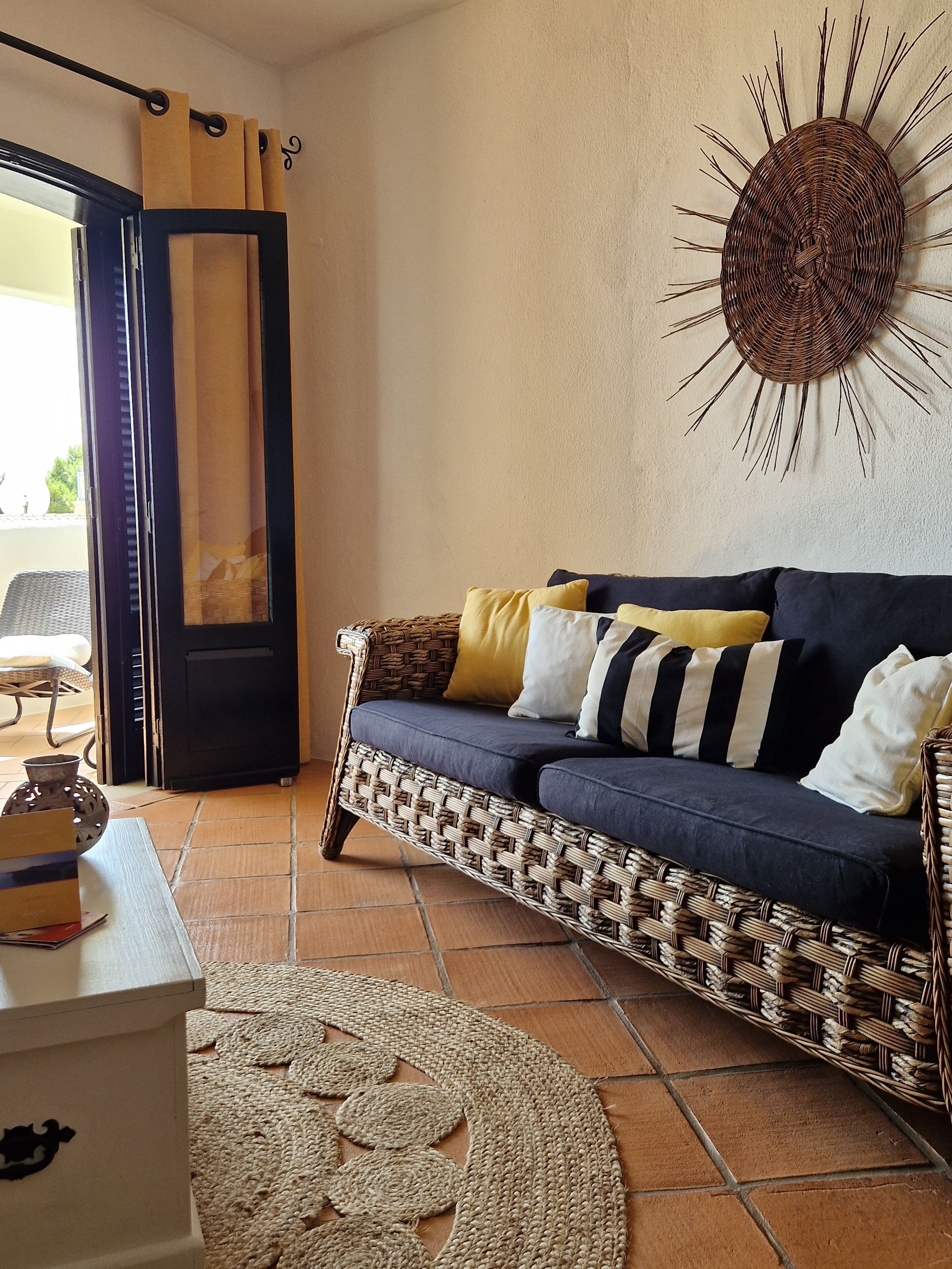 Una sala de estar con un sofá azul oscuro, almohadas amarillas y blancas, una mesa de centro tejida y un adorno de pared con diseño de sol.